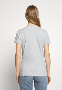 Žena s dlouhými blond vlasy, oblečená v jednoduchém světle šedém tričku s krátkým rukávem a modrých džínách, viděná zezadu na bílém pozadí.