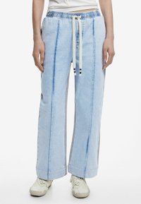 Pantalon en denim bleu clair à coupe large, doté d'une taille à cordon et de panneaux latéraux contrastants. Accompagné de chaussures claires.