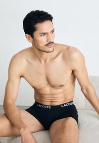 Black Lacoste boxer briefs with white waistband logo, soft fabric, fitted design, visible stitching, model seated on a light textured surface.