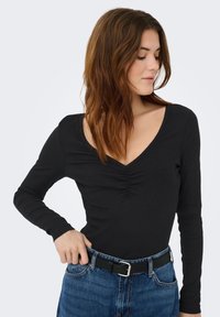 Sort langærmet top med V-hals og rynket frontdetalje, kombineret med højtaljede blå jeans og et sort bælte med sølvspænde.