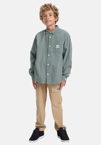 Groen en witte geblokte overhemd met knopen, beige cargobroek met zakken en zwarte sneakers. Bevat een logo-opdruk op het shirt.