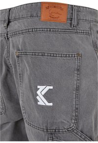 Pantalones de mezclilla gris con una etiqueta de cuero que lleva el texto "KARL KANI" y detalles del logo bordado en blanco en el bolsillo trasero. Acentos de herrajes metálicos.