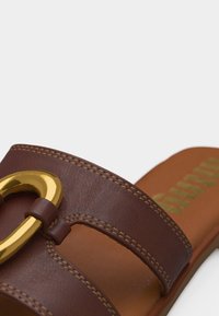 Sandale à glissière en cuir marron avec détail de boucle en métal doré et couture visible sur les brides, ainsi que semelle intérieure beige avec logo imprimé.