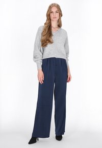 Grauer Strickpullover mit V-Ausschnitt und langen Ärmeln, kombiniert mit marineblauen weit geschnittenen Hosen. Das Modell trägt schwarze High Heels.