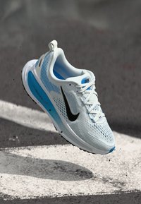 Kerge jooksu saabastega, millel on valgest ja sinisest õhuke ülemine osa, musta swoosh-logo, paelad ja tekstureeritud tald haardetamiseks.