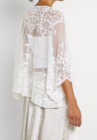 Person iført en gennemsigtig, hvid, blonde kimono-lignende top med blomsterbroderi over en hvid tube-top og en lysfarvet nederdel.