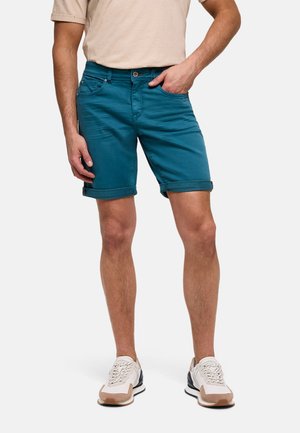 Hombre con pantalones cortos enrollados de color verde azulado, combinados con zapatillas blancas y beige y una camisa beige de manga corta, sobre un fondo liso.