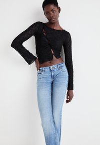 Jeune femme portant un crop top en dentelle noire à manches longues et un jean bleu clair, debout avec une main sur la hanche devant un fond blanc.