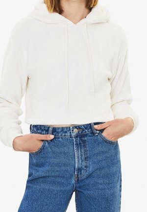 Personne portant un sweat à capuche blanc court avec cordons et un jean bleu taille haute, les mains reposant dans les poches avant.