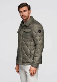 Uomo che indossa un giubbotto trapuntato verde oliva con tasche a patta sul petto e pantaloni bianchi, in piedi contro uno sfondo chiaro e uniforme.