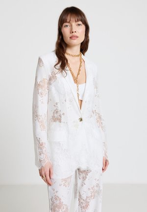 Frau trägt weißen Spitzenblazer und passende Hose, ergänzt durch mehrlagige goldene Ketten, steht vor einfarbigem Hintergrund.