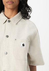 Beige short-sleeve shirt with a collared neckline, featuring a chest pocket and logo patch. Made of textured fabric with metallic buttons.