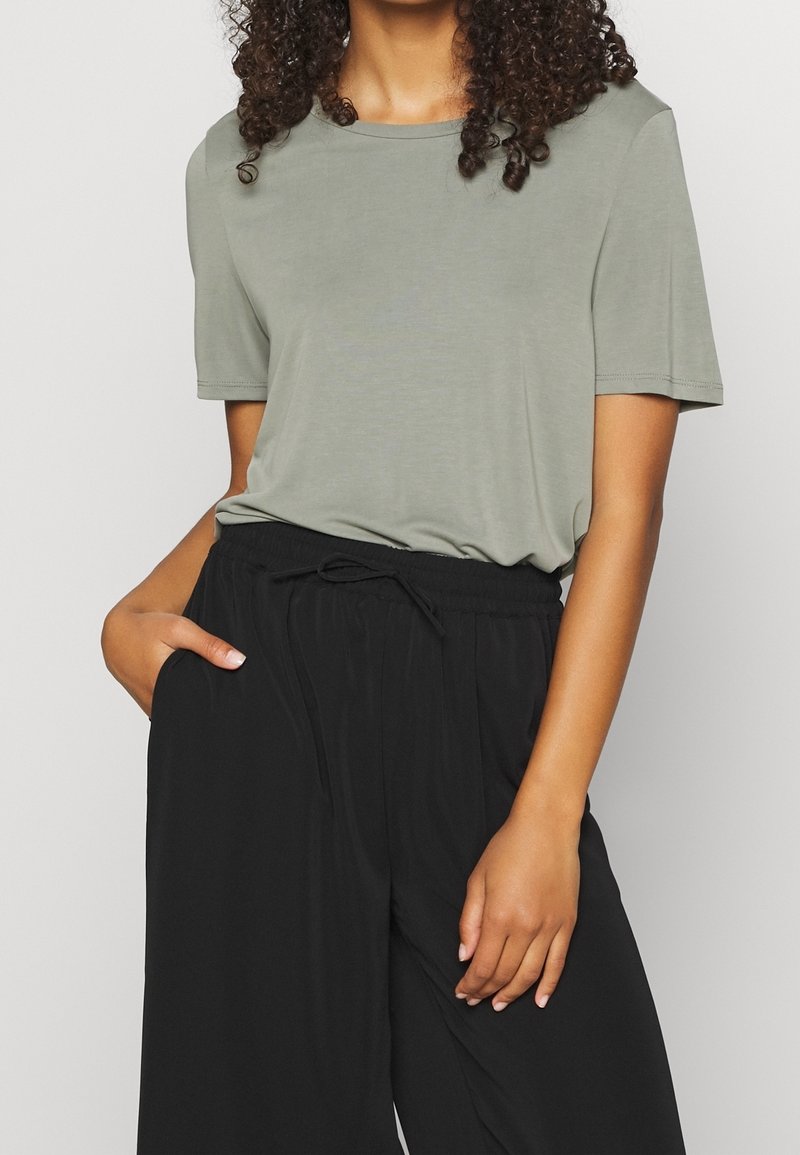 Femme portant une chemise à manches courtes vert clair rentrée dans un pantalon large noir avec une taille élastique et un cordon, main dans la poche.