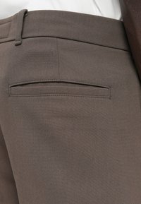 Pantaloni brown slim-fit realizzati in tessuto testurizzato, dotati di una tasca posteriore e passanti per la cintura. Finitura liscia con dettagli di cucitura discreti.