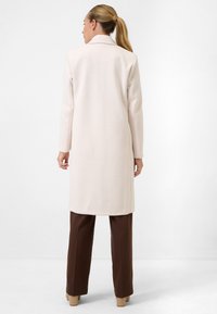 Lange beige jas met een gestructureerde afwerking, met een ingeponst kraag en lange mouwen, gecombineerd met een bruine broek en lichtgekleurde hakken.