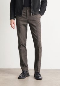 Pantaloni marroni, texturizzati, con una vestibilità sartoriale, dotati di chiusura a bottone e tasche laterali, abbinati a scarpe in pelle nera.