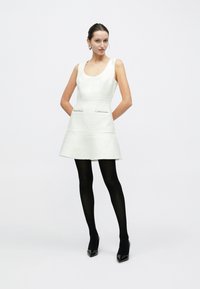 Biała sukienka o fakturze z okrągłym dekoltem i kieszeniami, ozdobiona metalicznymi akcentami. Modelka zestawia ją z czarnymi rajstopami i czółenkami na szpilce z lakierowanej skóry.