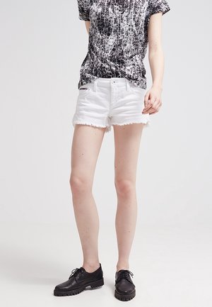 Persona che indossa una maglietta con stampa astratta bianca e nera, pantaloncini di jeans bianchi sfilacciati e scarpe nere con lacci, in piedi su uno sfondo semplice.