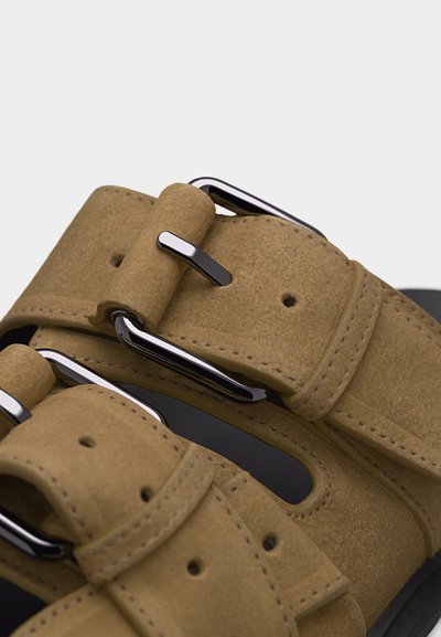 Κοντινή λήψη λουριών σανδαλιού από καφέ σουέντ με ασημένιες μεταλλικές αγκράφες και λεπτομερείς ραφές πάνω σε μαύρη σόλα.