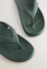 Paire de tongs en caoutchouc texturé vert foncé avec une bride en tige sur fond clair.