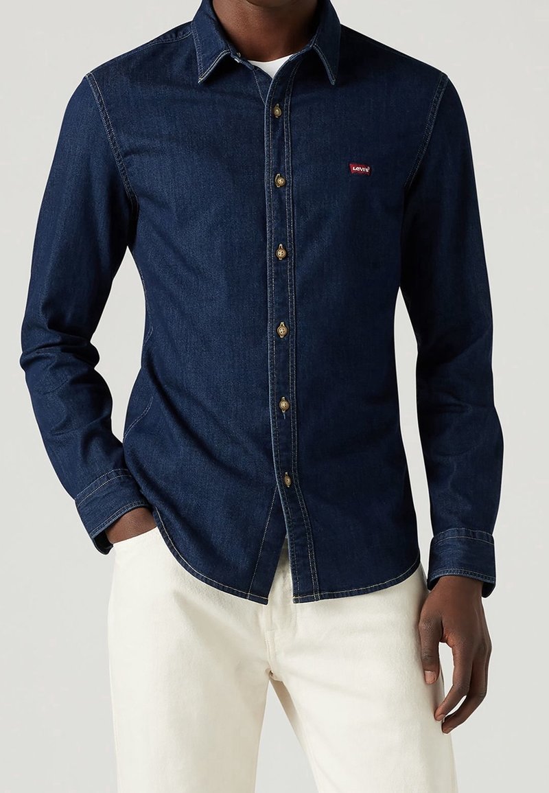 Homme portant une chemise en denim bleu foncé à boutons avec logo Levi’s et un pantalon blanc, debout avec une main dans la poche.
