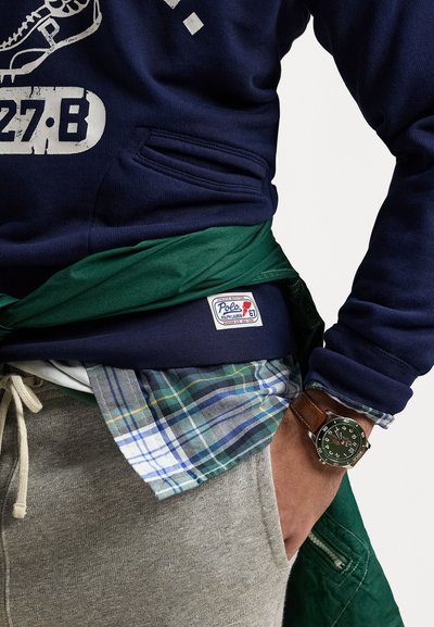 Hanorac bleumarin cu grafic alb, cămașă din flanel verde dedesubt, pantaloni de trening gri și un ceas din piele maro. Eticheta Polo pe hanorac.