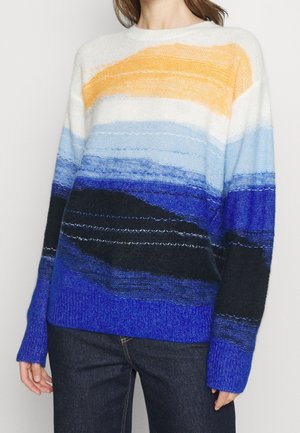 Femme portant un pull en maille multicolore à rayures horizontales jaunes, blanches, bleu clair, bleu foncé et noir, associé à un jean foncé.