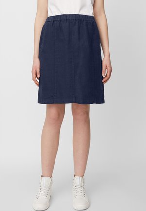 Femme portant une jupe marine jusqu'aux genoux avec ceinture élastique, un haut blanc et des baskets montantes blanches, se tenant contre un fond gris.