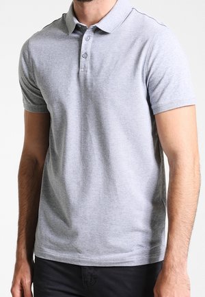 Mies, jolla on vaaleanharmaa lyhythihainen poolopaita, jossa on kolmen napin pystynappi ja mustat housut, yksinkertaista taustaa vasten.