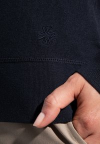 Granatowy sweter z dzianiny charakteryzujący się prążkowaną teksturą, haftowanym logo na dolnym brzegu oraz dopasowanymi mankietami. Ręka włożona do brązowej kieszeni poniżej.