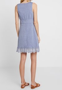 Blaues Chambray ärmelloses Kleid mit einem taillierten Oberteil, geraffter Taille und Spitzenbesatz am Saum. Getragen mit braunen Sandalen.