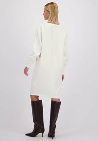 Witte, knieflokkerige jurk met lange mouwen en een relaxed fit; gestructureerde stof; gecombineerd met kniehoge, donkerbruine laarzen met hak.