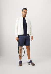 Mand iført hvid jakke, marineblå shorts og sneakers, der holder en tennisracket, stående mod en ensfarvet lys grå baggrund.