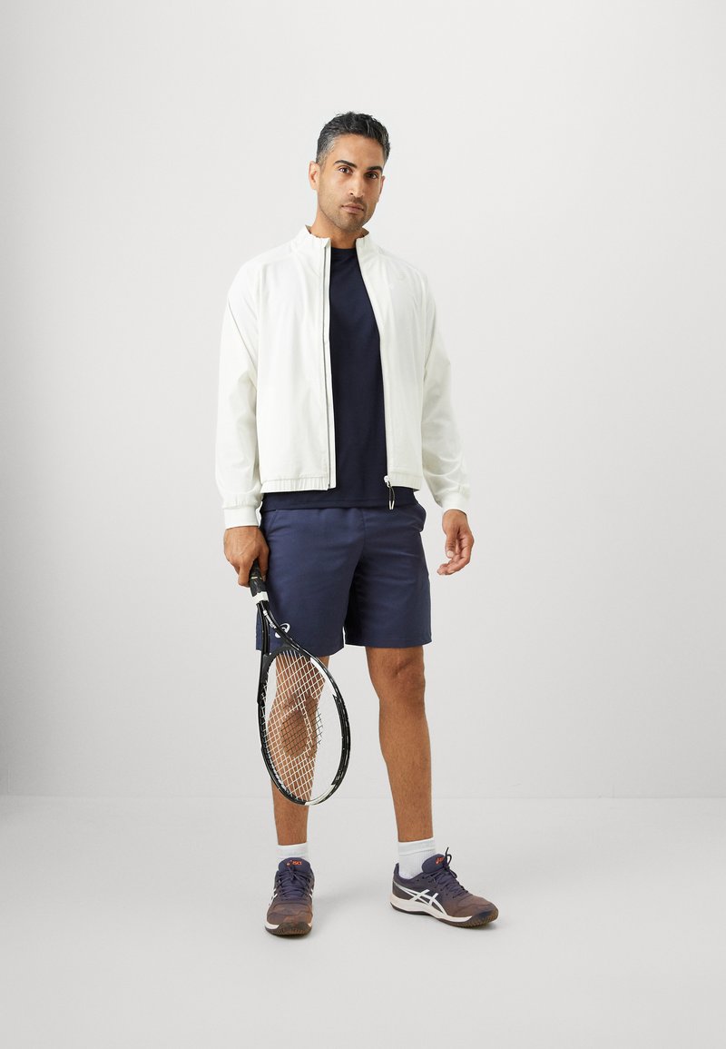 Mann in weißer Jacke, blauen Shorts und Turnschuhen, der einen Tennisschläger hält und vor einem einfarbigen hellgrauen Hintergrund steht.