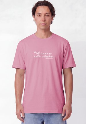 Mann mit mittellangen dunklen Haaren, der ein pinkes T-Shirt mit deutschem Text "Ich kann so nicht arbeiten" in weißer Handschrift trägt.