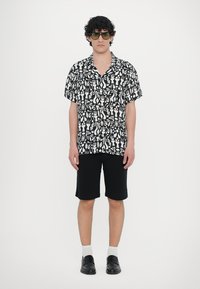 Κοντομάνικο πουκάμισο σε μαύρο και λευκό χρώμα με αφηρημένες σχεδιάσεις, συνδυασμένο με μαύρη βερμούδα, λευκές κάλτσες και μαύρα παπούτσια.