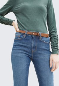 Farkut keskisinisessä sävyssä, joissa on viisi taskua, yhdistettynä ruskeaan nahkavyöhön, jossa on hopeinen solki, ja vihreään pitkähihalaiseen paitaan.