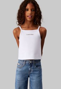 Hvit ermeløs crop-top med tynne stropper med "Calvin Klein"-logo i et subtilt trykk, kombinert med lyseblå denimjeans.