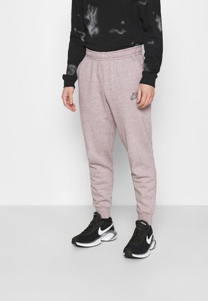 Persoon draagt lichtroze Nike joggingbroek en zwarte Nike sneakers met witte swoosh op een effen achtergrond.