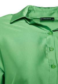 Grön satinblus med klassisk krage, knappslå framtill och slät textur; har matchande gröna knappar och en etikett på kragen.