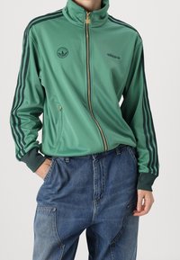 Grön polyester zip-up jacka med hög krage, marinblå ränder längs ärmarna, broderad logotyp på bröstet och sidofickor med dragkedja. Moderiktiga denimbyxor.