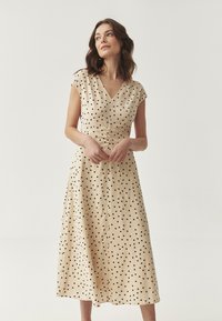 Beige midi-jurk met kapmouwen, V-hals en zwarte stippen. Beschikt over een gerimpelde taille en een vloeiende rok, gemaakt van lichte stof.