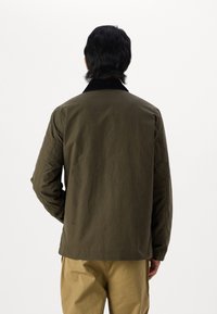 Giacca verde oliva realizzata in tessuto resistente, con colletto in velluto a coste nero e design a taglio dritto. Indossata sopra pantaloni khaki.