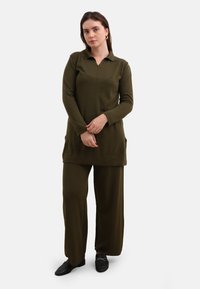 Olijfgroene set met lange mouwen, een tuniek met kraag en wijde broekspijpen. Zachte textuur, minimalistisch ontwerp, met zijzakken en eenvoudige stiksels.