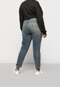 Osoba w jasnoniebieskich jeansach z wysokim stanem, czarnej bluzce z długim rękawem i białych butach Adidas, stojąca przodem do gładkiego, jasnego tła.