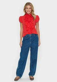 Blusa rossa a balze con maniche corte, abbinata a pantaloni in denim blu larghi e ballerine con stampa leopardata. Ensemble semplice e casual.