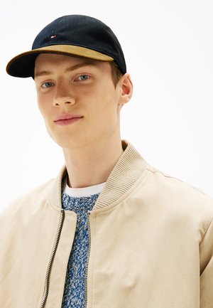 Black cap with brown brim, cotton material, featuring a small logo. Worn with a beige bomber jacket over a blue textured sweater.