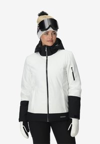Veste de ski blanche et noire avec fermeture éclair, deux poches latérales et une capuche noire. Le mannequin porte un bonnet beige et de grandes lunettes noires.