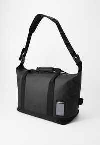 Svart duffelbag med teksturert nylonmateriale, justerbar skulderstropp, topphåndtak og et lite identifikasjonspanel på siden.