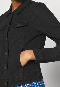 Czarna kurtka dżinsowa z kołnierzykiem, zatrzaskami, kieszeniami na piersi i dopasowanym krojem. Tkanina ma gładką teksturę.